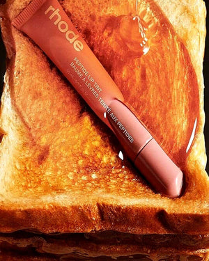 Rhode Retouch-Up Tint lip balm tube placed diagonally on a slice of toasted bread with a glossy drizzle of honey. The tube is a warm peachy-pink shade, and the background is the golden-brown toast.