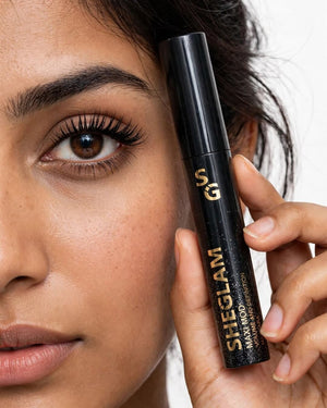 Pakistani model eye showing SHEGLAM Maxi Mod Volume and Definition Mascara result bold volumized defined fanned-out lashes with dense brush - holding enlarged real life black gold glitter tube