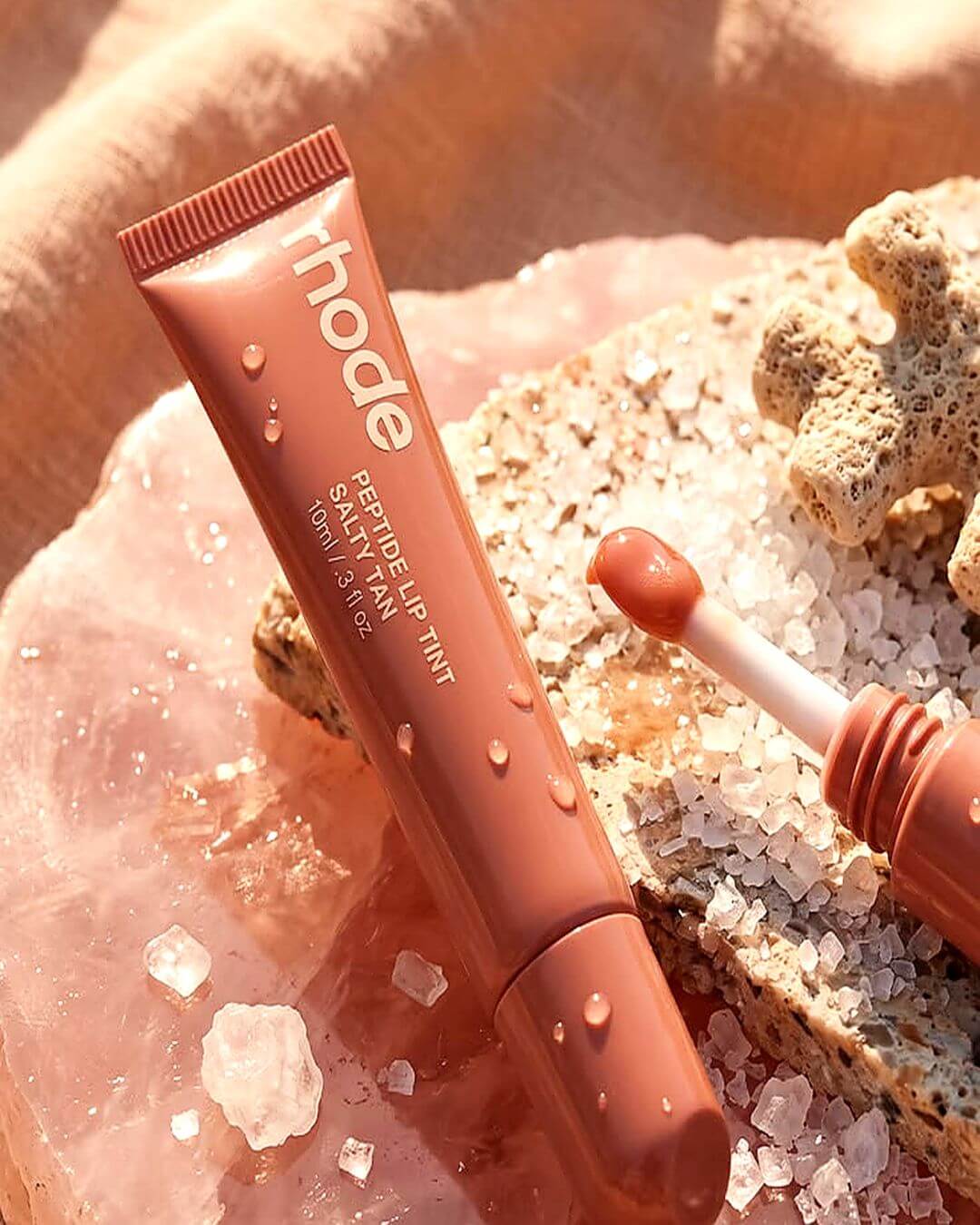 A tube of Thode Perpetual Lip Tint in a warm terracotta pink shade, styled on a pink crystal slab with salt crystals and coral against a linen backdrop.