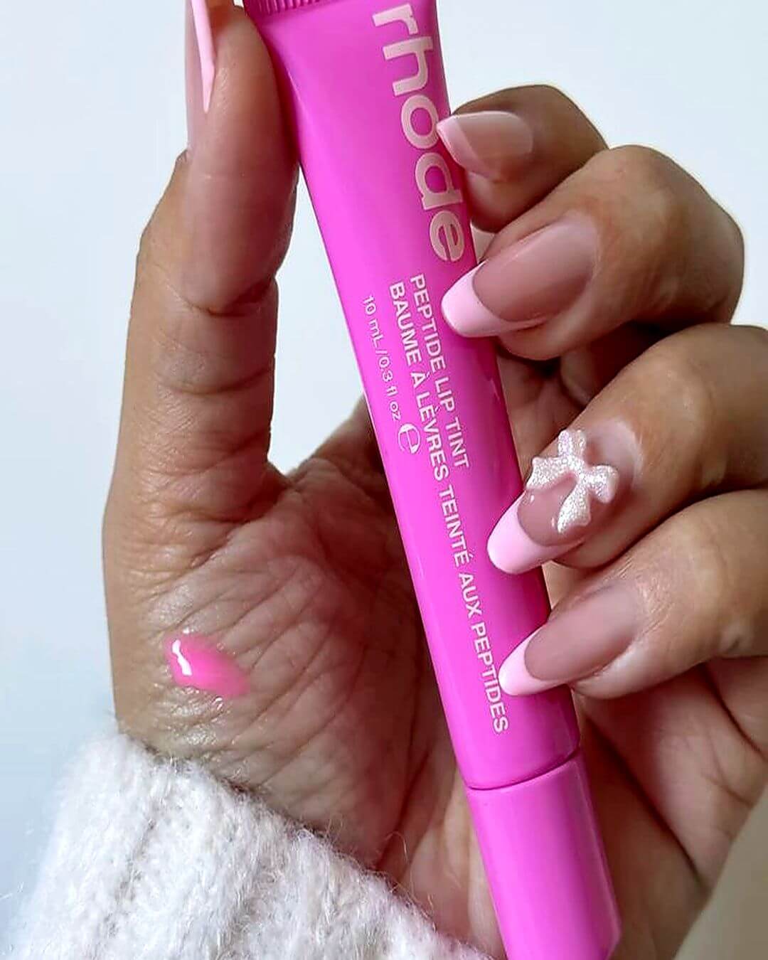 A close-up of a hand holding a bright pink tube of rhode Peptide Lip Tint Balm. The hand has a French manicure with one white glittery accent nail, and a small smear of the pink lip tint is visible on the skin.