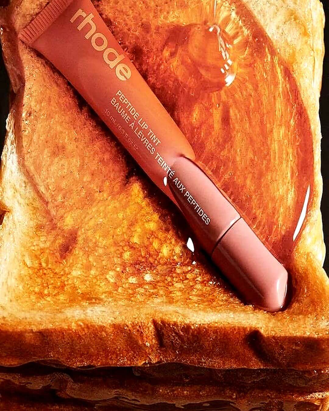 Rhode Retouch-Up Tint lip balm tube placed diagonally on a slice of toasted bread with a glossy drizzle of honey. The tube is a warm peachy-pink shade, and the background is the golden-brown toast.